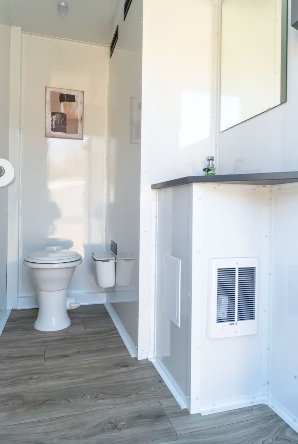 A clean, modern restroom fit for a Duchess or Duke, featuring a toilet, two waste bins, wall-mounted toilet paper, a sink with mirror, and elegant light wood flooring.