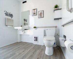 ADA-accessible bathroom with wooden floor, white walls, toilet, sink, handrails fit for a King & Queen, wall art, small plants, and a wall-mounted paper towel dispenser.