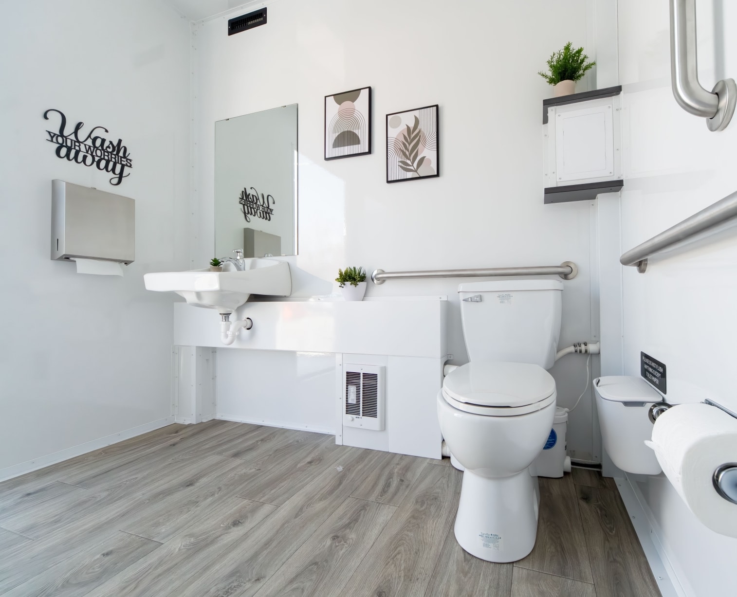 ADA-accessible bathroom with wooden floor, white walls, toilet, sink, handrails fit for a King & Queen, wall art, small plants, and a wall-mounted paper towel dispenser.