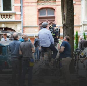 A film crew sets up a camera on a dolly track outdoors, with crew members standing nearby and historic buildings in the background.