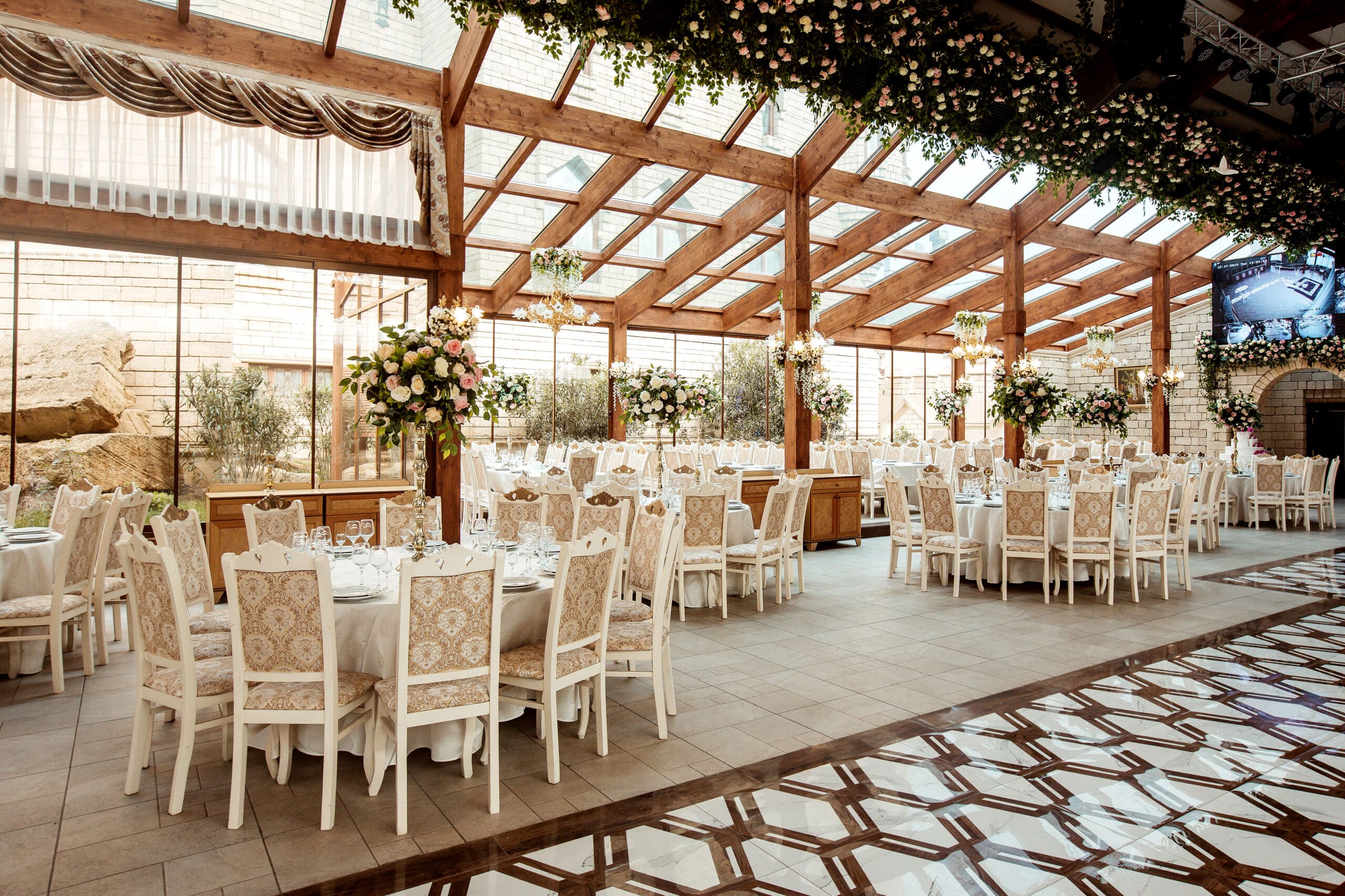 Elegant banquet hall with wooden beams, large windows, floral decorations, and round tables set for an event.