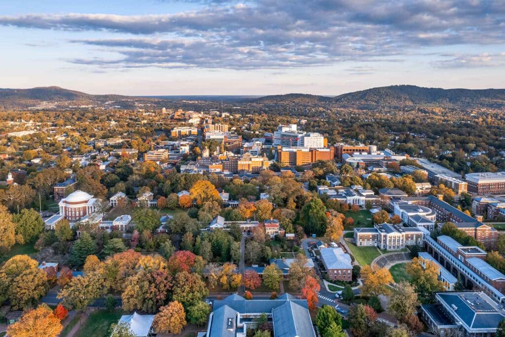 Aerial view of Charlottesville with clusters of buildings, tree-lined streets, and surrounding hills, taken on a clear day with scattered clouds.
