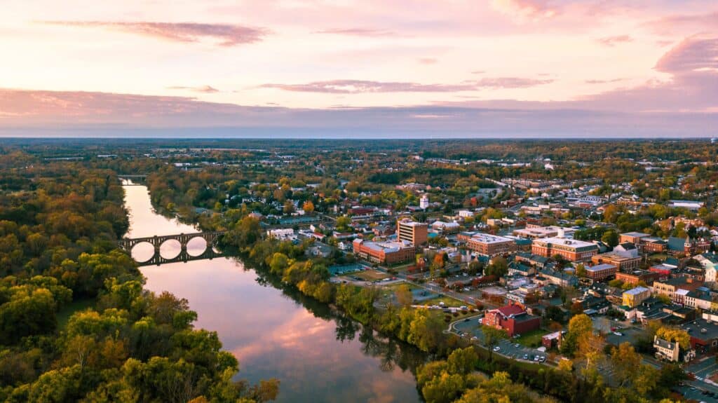 A river runs alongside Charlottesville with a large arched bridge, surrounded by trees and buildings under a colorful sunset sky.