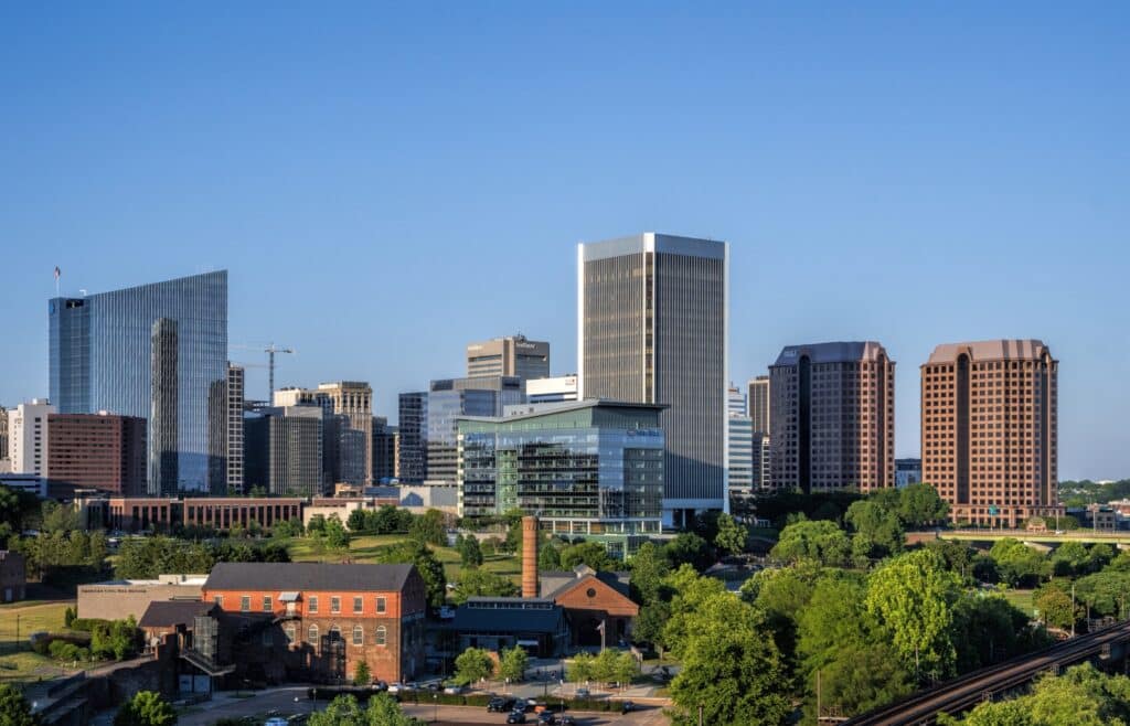 Skyline of Charlottesville with tall office buildings, historic brick structures, and green trees under a clear blue sky.