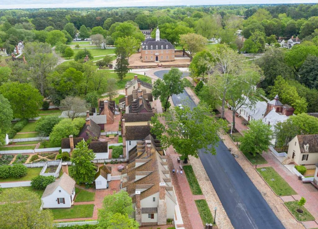 Aerial view of a historic colonial village in Charlottesville, with brick buildings, tree-lined streets, and manicured gardens, surrounded by greenery.