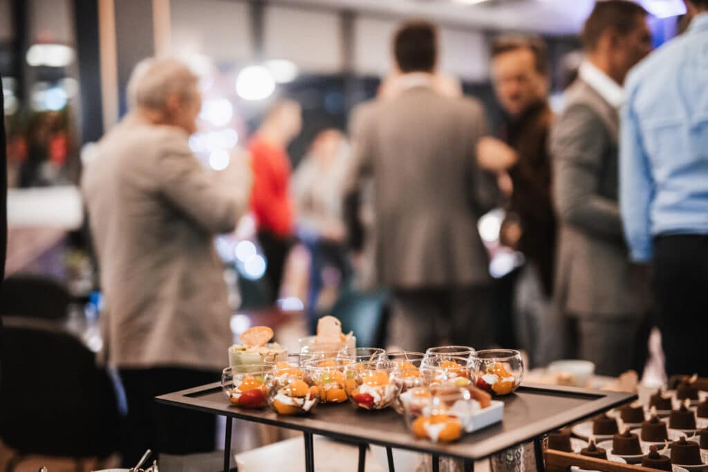 A table with desserts in glass cups in the foreground, perfect for Corporate Events, with a group of people in business attire socializing in the blurred background.