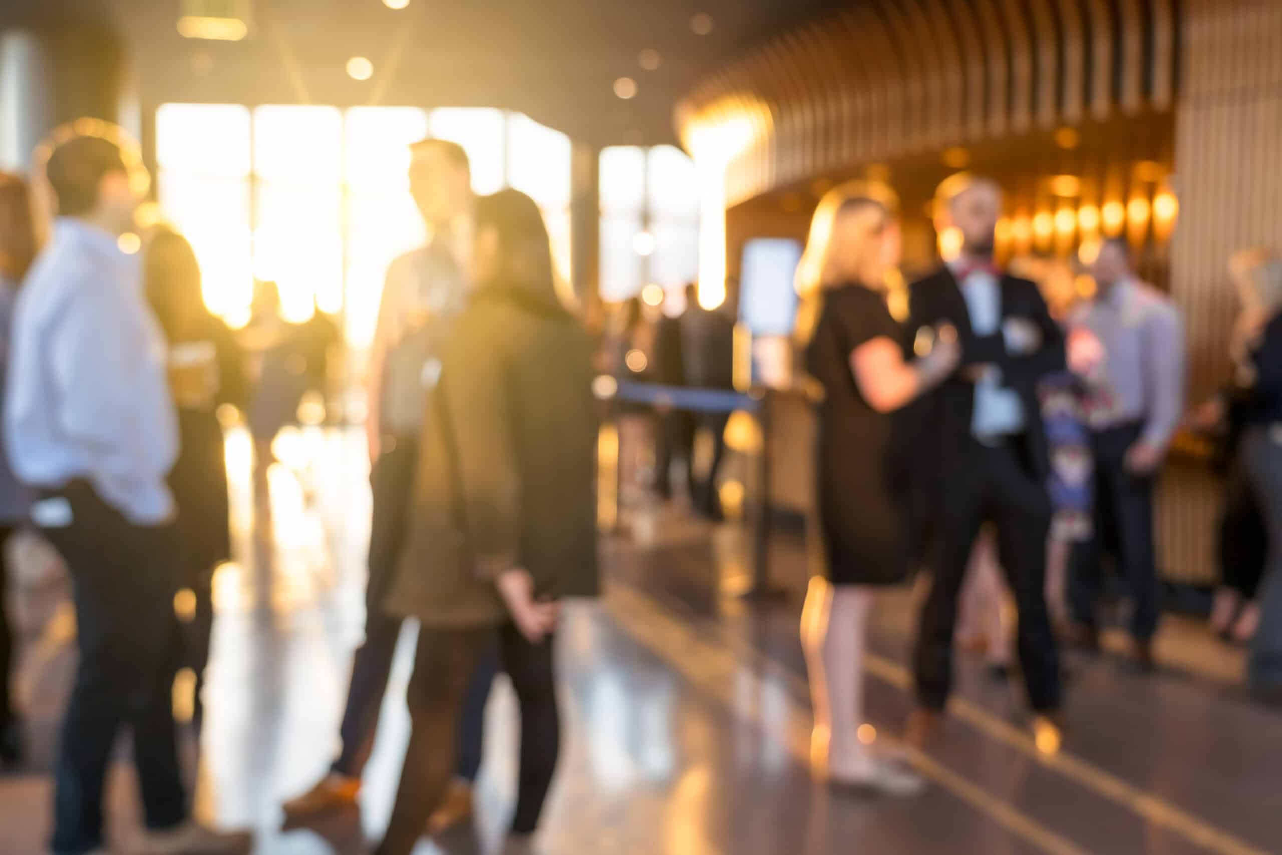 A group of people stand and talk in a modern, sunlit indoor space with large windows and blurred background, discussing ideas for corporate event planning.
