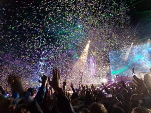 Concert audience with hands raised as confetti and lights fill the air, vibrant stage and performers in the background—festival comfort is elevated with luxury restroom trailers nearby.
