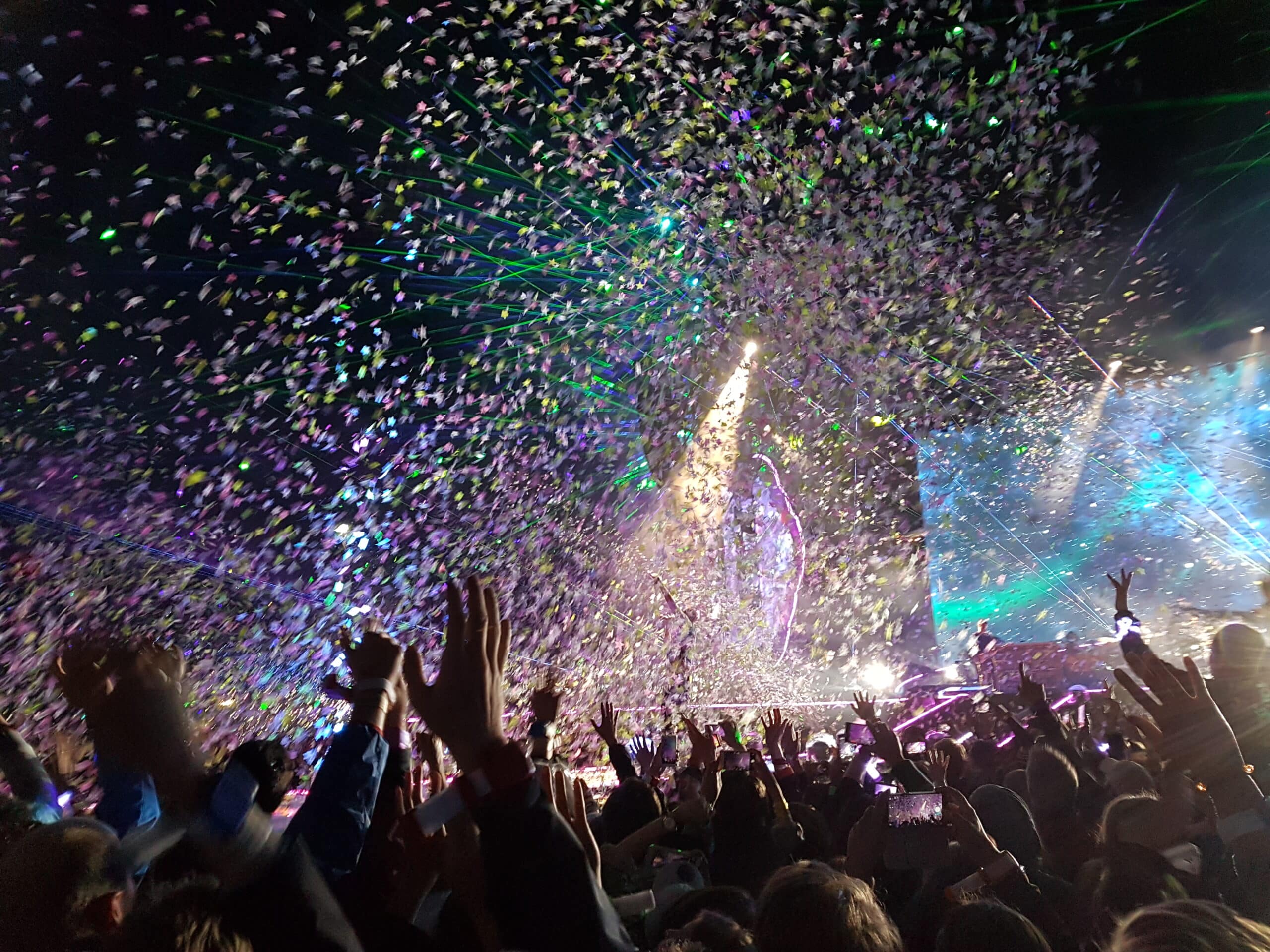 Concert audience with hands raised as confetti and lights fill the air, vibrant stage and performers in the background—festival comfort is elevated with luxury restroom trailers nearby.