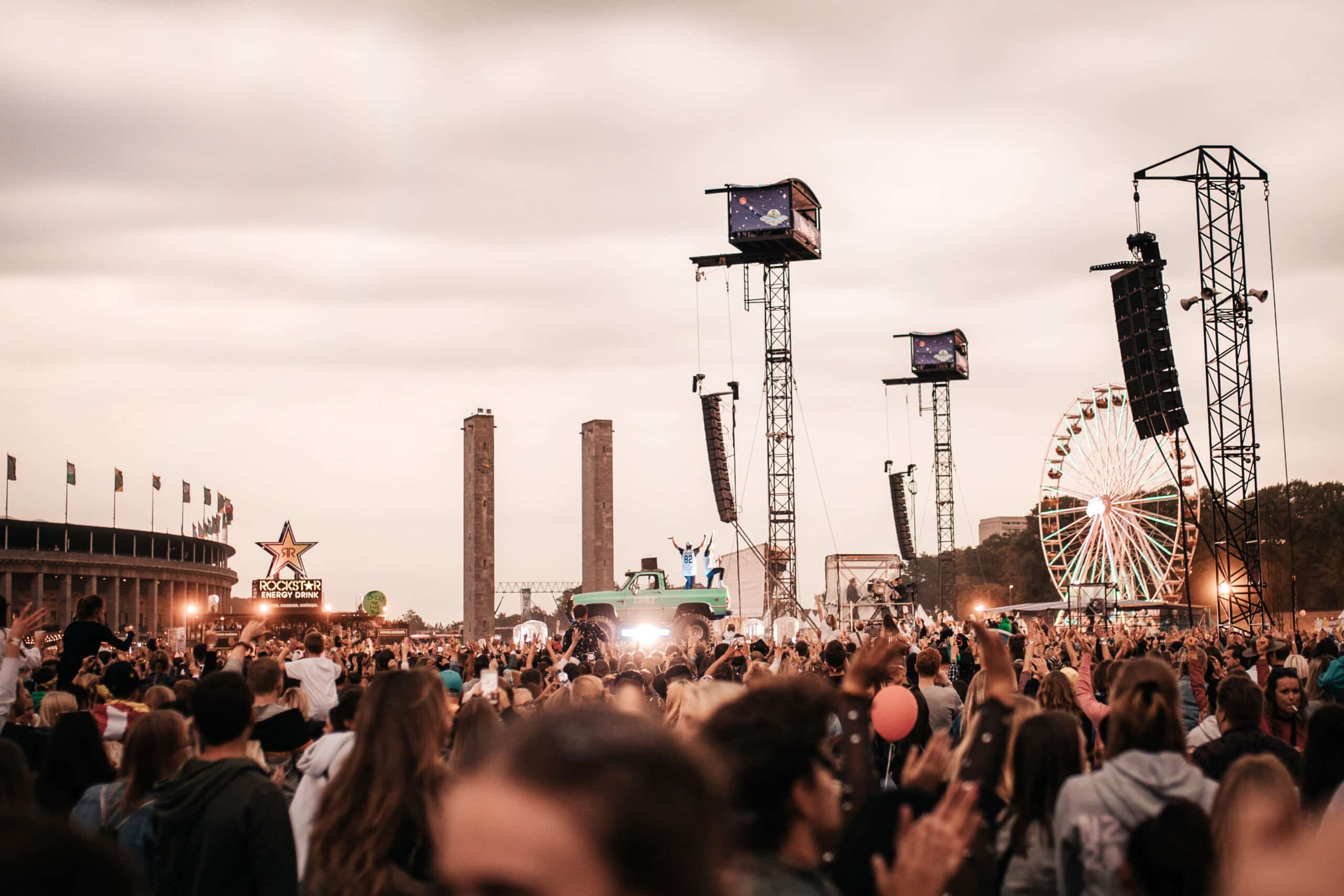 A large crowd of festival-goers gathers at an outdoor music festival, enjoying a vibrant festival experience with a performer on stage, a ferris wheel, and tall speaker towers set against a cloudy sky.