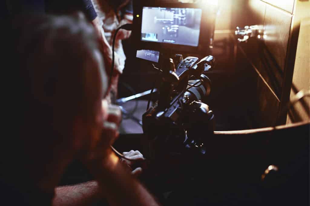 A person operates a professional video camera while monitoring footage on a screen in a dimly lit setting, showcasing the set logistics essential to smooth film production.