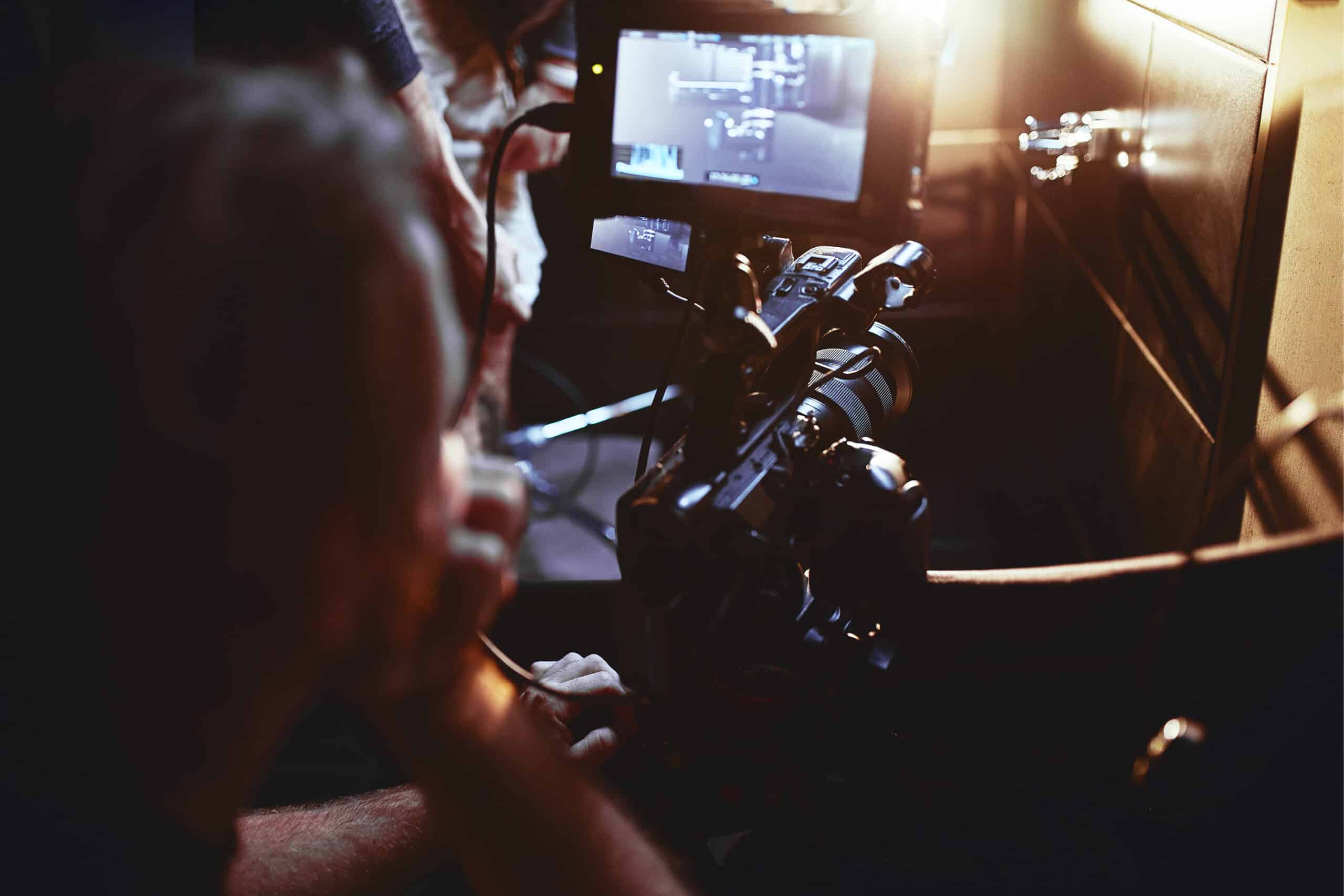 A person operates a professional video camera while monitoring footage on a screen in a dimly lit setting, showcasing the set logistics essential to smooth film production.