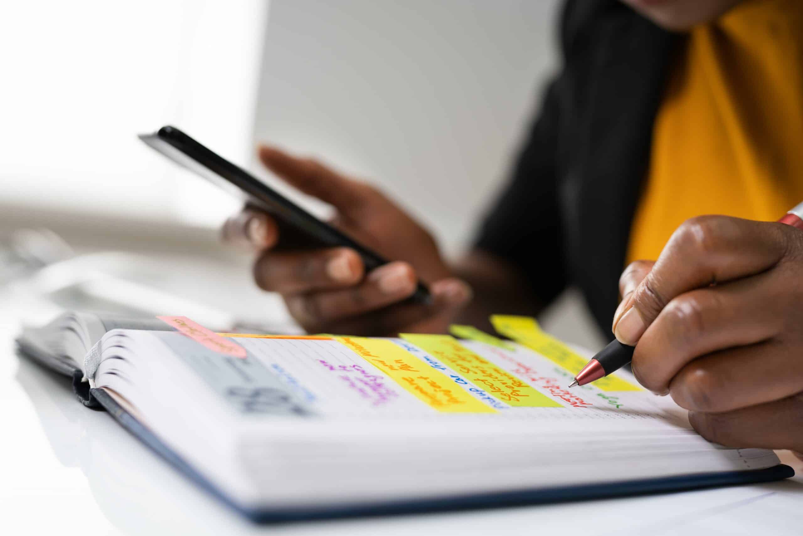 Person holding a smartphone and writing in a planner with colorful sticky notes and highlighted text, possibly organizing details for renting a restroom trailer.