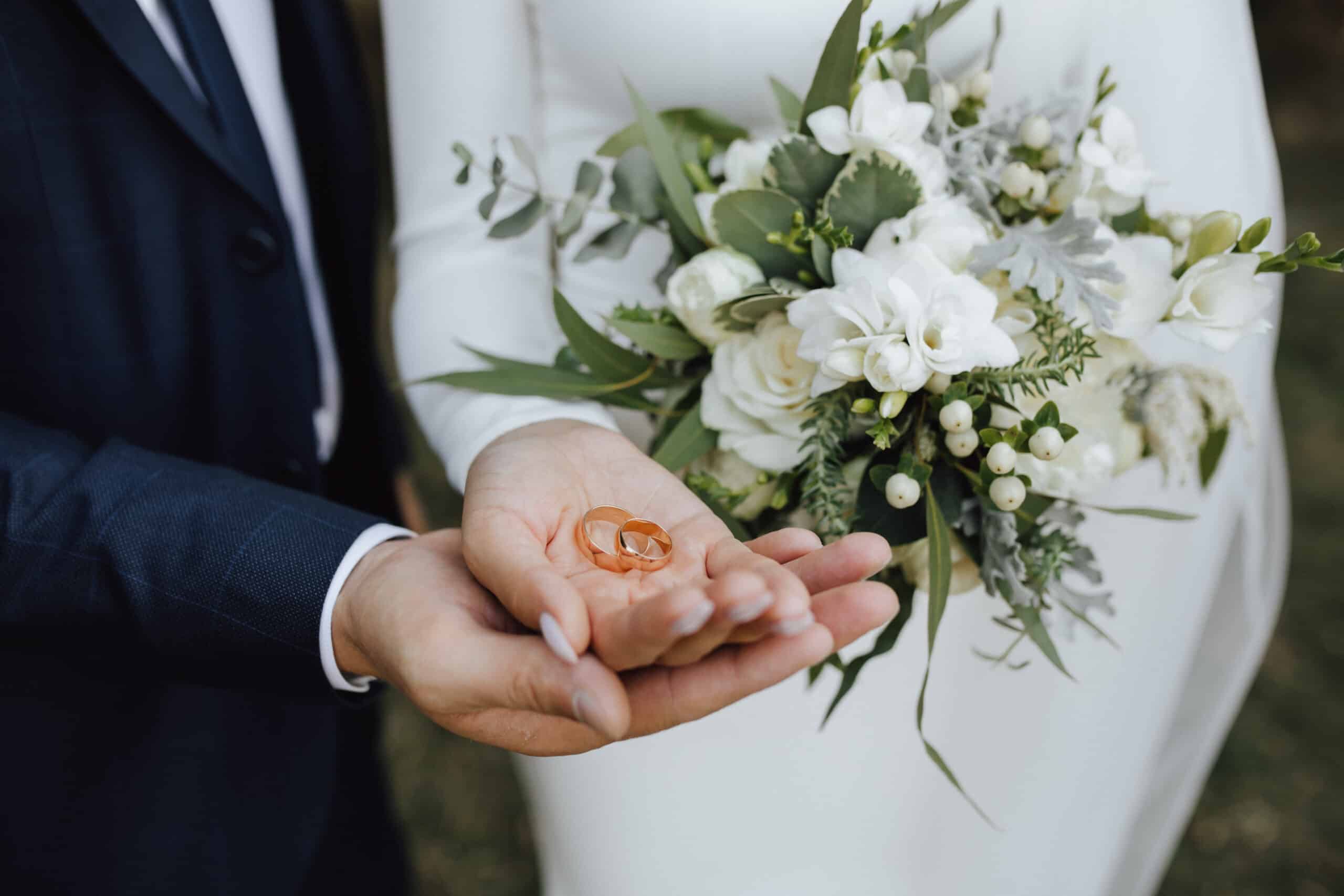 A person in a suit holds two gold wedding rings in their palm while another in a white dress, bouquet in hand, stands nearby—capturing the elegance often enhanced by luxury restroom trailers at upscale weddings.